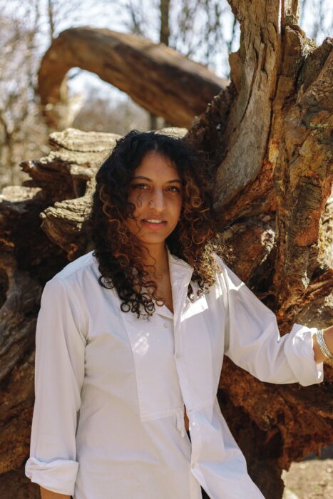 Sheena Patel framför en majestätisk trädrot, vilket återspeglar hennes djärva närvaro i litteraturvärlden med sin debutroman "Jag är ett fan".