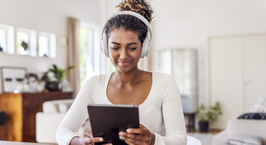 En ung kvinna sitter vid ett bord och tittar på sin läsplatta. Hon har hörlurar på huvudet. Hon har mörkt, uppsatt hår.